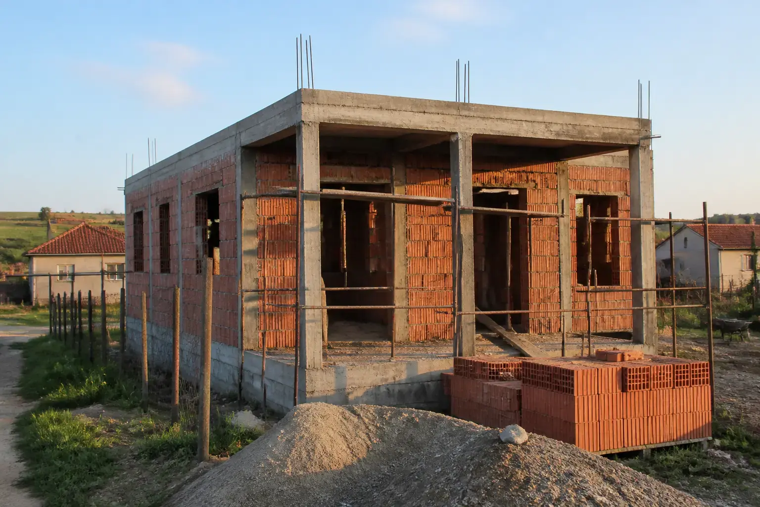O casă aflată în stadiul de construcție la roșu, realizată din cărămidă și beton, situată într-o zonă rurală cu alte case vizibile în fundal